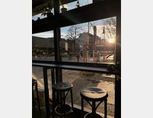 Blick aus einem Café mit großen Glasfenstern auf eine Straße bei tiefstehender Sonne. Im Vordergrund stehen Barhocker, draußen sind Fußgänger, ein Fahrradhalter und Bäume sichtbar, mit Gebäuden im Hintergrund.