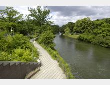 Uferweg entlang eines Flusses mit üppiger grüner Vegetation, gepflastertem Fußweg und schmiedeeisernem Geländer. Links befinden sich eine Backsteinmauer mit Treppe und Straßenlaternen im klassischen Stil.