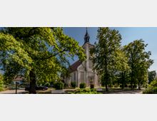 Kirche in einem parkähnlichen Umfeld mit großen, belaubten Bäumen und Bänken im Vordergrund. Das Gebäude hat eine helle Fassade, ein rotes Dach und einen Kirchturm mit einer Spitze, die vor dem blauen Himmel hervorragt.