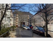 Mehrstöckige Wohnhäuser mit beigefarbenen Fassaden und Holzfensterläden bilden einen Innenhof. Vor den Gebäuden stehen parkende Autos, Bäume ohne Laub und ein gepflasterter Gehweg säumen die Straße.