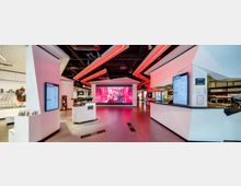 Moderner Innenraum eines multifunktionalen Ladens oder Erlebniszentrums mit hellen Wänden, roten LED-Deckenleuchten und mehreren Bildschirmen. Im Vordergrund sind weiße Info- und Verkaufstheken zu sehen, im Hintergrund eine große digitale Leinwand und ein Bereich mit Regalen sowie gastronomischen Angeboten.