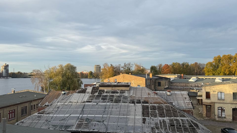Blick über ein ehemaliges Industrie- oder Lagerareal mit flachen Ziegel- und Blechdächern, teils verwittert und mit Abdichtbahnen bedeckt. Im Hintergrund liegen ein breiter Fluss, eine Brücke, vereinzelte Hochhäuser sowie herbstliche Bäume unter einem bewölkten Himmel.