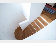 Holztreppe in einem modernen Innenraum, die sich in einer geschwungenen Form nach unten windet, mit weißen Wänden und einem Holzboden im oberen Bereich. Im Hintergrund ist ein bunter Teppich sichtbar.