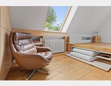 Gemütlicher Dachraum mit Holzfußboden, einer braunen Ledersessel auf einem Metallgestell, einem eingebauten Regal mit Büchern und einem Heizkörper unter einem großen Dachfenster. Im Vordergrund ein multifunktionales Holzpodest mit herausziehbaren Matratzen.