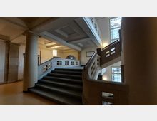 Innenraum eines historischen Gebäudes mit einer breiten, geschwungenen Treppe aus Stein, verziert mit Geländern und klassischen Säulen. An den Wänden befinden sich große Fenster mit buntem Glas und dekorative Jugendstil-Elemente, die von warmem Licht beleuchtet werden.