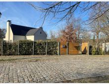 Ein modernes Wohnhaus mit hellem Putz und schwarzem Satteldach, umgeben von einer Hecke und einem Zaun mit Holztoren. Im Vordergrund befindet sich eine gepflasterte Straße, und im Hintergrund sind kahle Bäume unter klarem Himmel zu sehen.