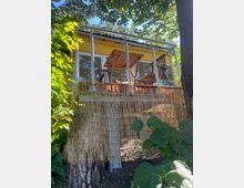 Gelbes Gartenhaus mit erhöhtem Balkon, ausgestattet mit Holzstühlen und einem Tisch, umgeben von dichter Vegetation. Unterhalb des Balkons ist eine Bambusmatte als Sichtschutz angebracht.