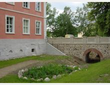 Rosa gestrichenes Gebäude mit weißen Fensterrahmen, daneben eine Steinbrücke mit einem kleinen gemauerten Rundbogen. Der Vordergrund zeigt eine Rasenfläche mit Blumenbeet, umgeben von Steinen. Im Hintergrund sind Bäume zu sehen.