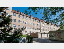 Mehrstöckiges Wohngebäude mit einfacher Fassade und mehreren weißen Fenstern, davor eine Reihe von Garagen mit grauen Toren und graffitibeschmierte Wände. Im Vordergrund sind Bäume und Sträucher zu sehen, der Himmel ist klar und blau.