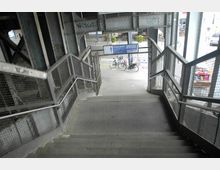 Treppe unter einer metallenen Brückenkonstruktion mit seitlichen Geländern aus Drahtgitter führt hinab zu einem Gehweg. Im Hintergrund sind Fahrräder, Schilder und ein Straßenbereich zu sehen.