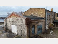 Verwitterter Backstein-Industriebau mit flachem Dach und Anbauten, teils verputzter Giebelwand und großen Tor- und Fensteröffnungen. Im Vordergrund liegt ein unbefestigter Hof mit überwucherten Bereichen; im Hintergrund ist ein Gewässer neben dem Gebäudekomplex zu sehen.