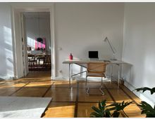 Helles Zimmer mit weiß gestrichenen Wänden und Parkettboden. Im Vordergrund steht ein Schreibtisch mit Laptop, Lampe und einem Stuhl aus Holz und Metall; im Hintergrund ein angrenzendes Esszimmer mit hellem Holztisch, Stühlen und einem pinken Kunstwerk an der Wand.