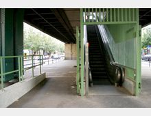 Eine Rolltreppe unter einer erhöhten Bahnbrücke führt nach oben, umgeben von grün gestrichenen Metallgittern. Im Hintergrund sind Bürgersteige, geparkte Autos und eine belebte Straße mit Bäumen sichtbar.