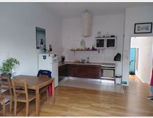 Wohnküche mit Holzboden und weiß gestrichenen Wänden. Links steht ein Esstisch aus Holz mit Stühlen, rechts eine Küchenzeile mit dunklen Schränken, weiß gefliester Rückwand, Herd und Hängeschränken; im Hintergrund ein Kühlschrank und eine offene Tür zu einem Flur.