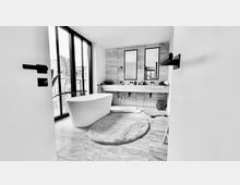 Schwarz-weiß-Aufnahme eines modernen Badezimmers mit freistehender Badewanne, großen bodentiefen Fenstern und einer Wand mit zwei rechteckigen Spiegeln über einem breiten Waschtisch. Der Boden besteht aus hellen Fliesen, und ein runder Teppich liegt zentral im Raum.