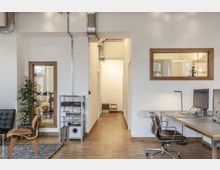 Modernes Büro mit minimalistischem Design, Holzboden und weißen Wänden. Das Bild zeigt einen Arbeitsbereich mit Schreibtischen, Computern und Bürostühlen sowie einer schmalen Durchgangstür zu einem Flur; rechts und links befinden sich Fenster mit Holzrahmen.