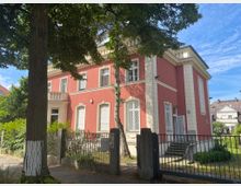 Zweistöckiges Gebäude mit rot gestrichener Fassade und weißen Fensterrahmen, flankiert von hellen Zierelementen. Vor dem Haus befindet sich ein Metallzaun mit Tor, dahinter ein kleiner Vorgarten mit Bäumen und Sträuchern.
