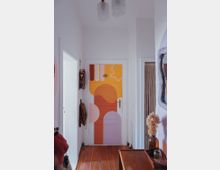 Ein schmaler Flur in einer Wohnung mit Holzboden und weißen Wänden, während die Tür am Ende des Flurs farbenfroh mit abstrakten geometrischen Mustern in Lila, Orange und Gelb bemalt ist. Links an der Wand hängt ein Rucksack, und rechts befindet sich ein kleiner Tisch mit einer Vase und getrockneten Blumen.