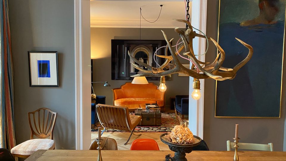 Blick von einem Essbereich mit langem Holztisch und Kerzenhaltern durch eine breite, weiß gerahmte Türöffnung in ein angrenzendes Wohnzimmer. Im Wohnzimmer stehen ein orangefarbenes Sofa, dunkle Sessel und ein Couchtisch auf einem gemusterten Teppich; im Vordergrund hängt eine Geweihleuchte mit freiliegenden Glühbirnen, und an den grauen Wänden hängen gerahmte Bilder.