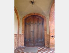 Holztür mit dekorativen schwarzen Beschlägen in einem Eingangsbogen aus roten Ziegelsteinen. Die Decke über der Tür ist beige, verziert mit floralen Elementen und einer zentralen Lampe.