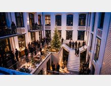 Innenhof eines mehrstöckigen Gebäudes mit hohen, bogenförmigen Fenstern und umlaufenden Galerien. In der Mitte steht ein beleuchteter Weihnachtsbaum; Treppen, Glasgeländer, Laternen und schneebedeckte Dekorationen prägen die winterliche Atmosphäre.