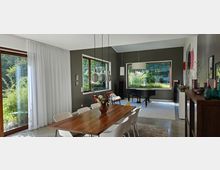 Helles Wohnzimmer mit Essbereich, einem großen Holztisch und weißen Stühlen. Große Fenster mit Blick in einen grünen Garten, ein schwarzer Flügel auf einem Teppich und moderne Hängelampen dominieren den Raum, der durch schlichte Deko und klare Linien geprägt ist.