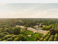 Luftaufnahme eines historischen Parks mit einem barocken Gebäude im Vordergrund, umgeben von symmetrischen Gartenanlagen und Bäumen. Im Hintergrund sind eine historische Mühle und ein weiteres Schlossgebäude zu sehen, eingebettet in eine waldreiche Landschaft.