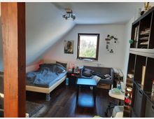 Dachgeschosszimmer mit schräger Decke, ausgestattet mit einem Bett, einem kleinen Sofa, einem schwarzen Couchtisch und einem großen Regal rechts im Bild. Ein kleines Fenster bietet Blick auf eine grüne Landschaft, der Fußboden ist aus dunklem Holz.