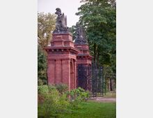 Rotes Backsteinportal mit dekorativen Skulpturen und verzierten schwarzen Metalltoren in einem parkähnlichen Umfeld, umgeben von Bäumen und Rasenflächen.