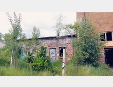 Verlassene Backsteinruine mit teilweise zerstörten Wänden, überwuchert mit jungen Bäumen und Sträuchern; im Vordergrund eine grüne Wiese, im Hintergrund ein höheres Gebäude mit offenen Fenstern.