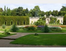 Ein gepflegter Schlossgarten mit symmetrischen Blumenbeeten in leuchtenden Farben, Rasenflächen und geschwungenen Wegen. Im Hintergrund eine Steinbalustrade mit Statuen und ein Tor, umgeben von dichtem Grün.