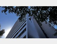 Hochhausfassade aus Glas und Metall, aufgenommen aus einem niedrigen Winkel gegen einen klaren blauen Himmel; im oberen Bereich ragen Baumäste ins Bild.