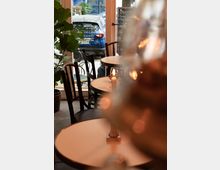 Innenraum eines kleinen Cafés mit runden Tischen und dunklen Holzstühlen, einer großen grünen Pflanze und einer Glasfassade, die den Blick auf ein geparktes blaues Auto und eine Straße ermöglicht. Auf den Tischen stehen brennende Kerzen in metallenen Haltern.