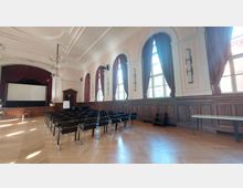 Ein klassischer Veranstaltungsraum mit Parkettboden, hohen Fenstern mit schweren, weinroten Vorhängen und einer Bühne mit einem weißen Projektionsschirm. Die Wände sind mit Holzvertäfelungen und dekorativen Stuckdetails gestaltet, und Reihen von schwarzen Stühlen sind vor der Bühne aufgestellt.