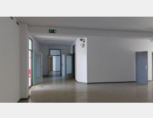 Leerer Flur in einem Büro- oder Verwaltungsgebäude mit weißen Wänden und grauem Boden. Mehrere geöffnete Türen aus Glas und Holz führen in angrenzende Räume, oben befindet sich ein Notausgangsschild.