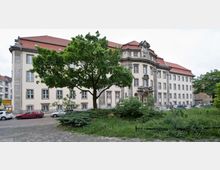 Historisches Verwaltungsgebäude mit heller Fassade, symmetrischen Fenstern und einem zentralen Eingangsbereich mit kunstvollen Steinverzierungen. Im Vordergrund befinden sich ein Baum, angelegte Grünflächen und parkende Autos auf einer gepflasterten Straße.