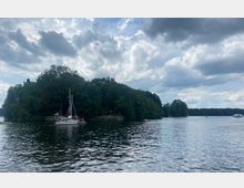 Waldreiche Uferlandschaft an einem See mit einem Segelboot im Vordergrund und weiteren Booten im Hintergrund unter wolkenbedecktem Himmel.