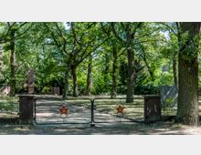 Eingang zu einem Friedhof oder Gedenkort mit Metalltor, das rote Symbole von Hammer und Sichel enthält. Im Hintergrund sind hohe Bäume, Pfade und monumentale Stele-Strukturen in einem parkähnlichen Umfeld zu sehen.