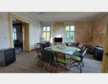 Ein einfacher, renovierungsbedürftiger Raum mit Holzboden, abblätterndem Wandputz und hohen Fenstern, die Tageslicht hereinlassen. In der Mitte steht ein rechteckiger Tisch mit Stühlen, gedeckt mit einer Tischdecke und Getränken; im Hintergrund stehen ein Fernseher und Möbel.