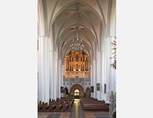 Innenansicht einer hohen gotischen Kirche mit spitzbogigen Gewölben, weißen Pfeilern und einer prächtigen, goldverzierten Orgel auf einer Empore im Hintergrund. Darunter befinden sich ein Rundbogenportal und Kirchenbänke, die den Mittelgang flankieren.