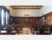 Rats- oder Sitzungssaal mit hölzernen Wänden und Möbeln, darunter Reihen von Stühlen und Tischen mit hohen Rückenlehnen. Im Hintergrund befinden sich ein Gemälde, mehrere Flaggen sowie eine große, zentrale Hängelampe an einer Kassettendecke. Links sind hohe Fenster mit farbigem Glas zu sehen.