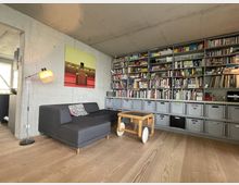 Wohnzimmer mit moderner Einrichtung, bestehend aus einem grauen Sofa, einem Holz-Tisch auf Rädern, einer Stehlampe und einer großen, wandfüllenden Regalwand mit Büchern und Aufbewahrungsboxen. Die Wände sind aus Sichtbeton, der Boden ist aus hellem Holz.