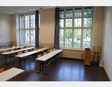 Ein Klassenraum mit mehreren weißen Tischen und Holzstühlen, angeordnet in Reihen. Große Fenster mit Gittermustern lassen Tageslicht herein; dahinter sind grüne Büsche und parkende Autos sichtbar.