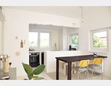 Helle, minimalistisch eingerichtete Wohnküche mit weiß gestrichenen Wänden, großem Esstisch aus dunklem Holz, gelben Stühlen und einer offenen Durchreiche zur Küche. Große Fenster lassen Tageslicht herein, und draußen sind grüne Bäume sichtbar.