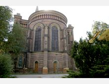 Außenansicht einer historischen Backsteinkirche mit halbrunder Apsis, großen gotischen Fenstern und dekorativen Bögen. Im Vordergrund befinden sich Bäume und Sträucher, die teilweise die Gebäudeansicht umrahmen.