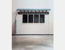 Schlichter Innenraum mit einem Wandabschnitt, an dem eine schwarze Tafel mit der Aufschrift „VORANKÜNDIGUNGEN“ sowie vier leere Schaukästen befestigt sind. Der Boden besteht aus glattem, grauem Beton, und die Wand ist weiß gestrichen.