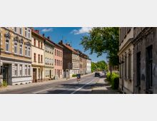 Straßenszene mit Reihen von mehrstöckigen Altbauten in unterschiedlichen Farben auf der linken Seite und einem Gehweg mit Bäumen auf der rechten Seite. Die Straße ist mäßig befahren, mit einem geparkten Auto und einer Baustellenabsperrung im Vordergrund.