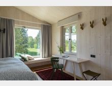 Holzvertäfeltes Schlafzimmer mit einem großen Fenster, das den Blick auf eine grüne Wiese und Bäume freigibt. Im Raum befinden sich ein Schreibtisch mit einem Stuhl, ein Bett mit einer grauen Decke und eine Fensterbank mit Kissen.