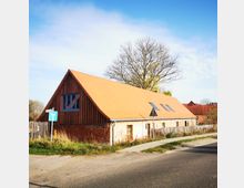 Backsteinhaus mit Satteldach aus orangenen Ziegeln, blau gestrichenen Fenstern und Holzverkleidung im oberen Bereich. Vor dem Gebäude stehen ein Verkehrszeichen und ein kahler Baum; daneben ein Gehweg und ein Zaun.