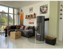 Wohnzimmer mit großen bodentiefen Fenstern, grauem Ecksofa und bunten Kissen. Ein Kaminofen steht an der Wand, die mit Kunst und Dekorationen geschmückt ist, und ein heller Holzboden reflektiert das Licht.